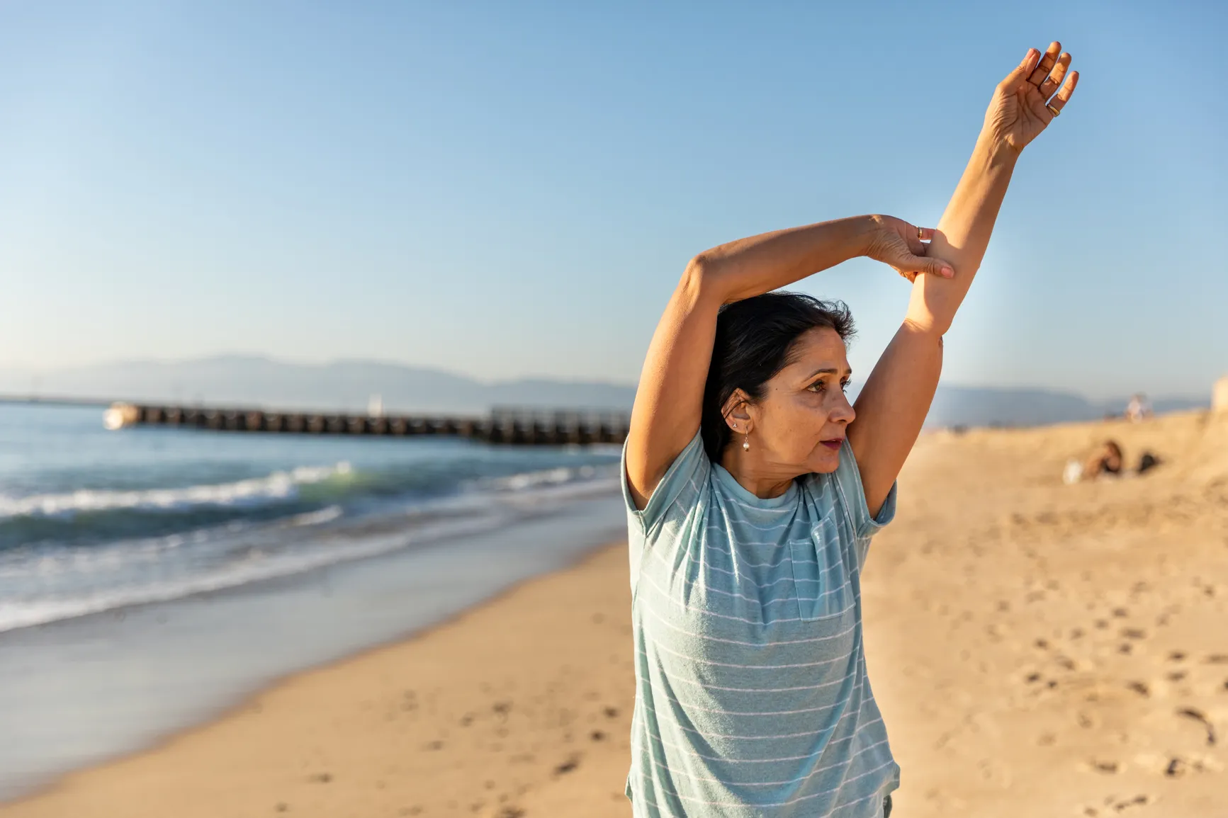 Woman on the beach wearing a striped t-shirt gently stretches her arms overhead